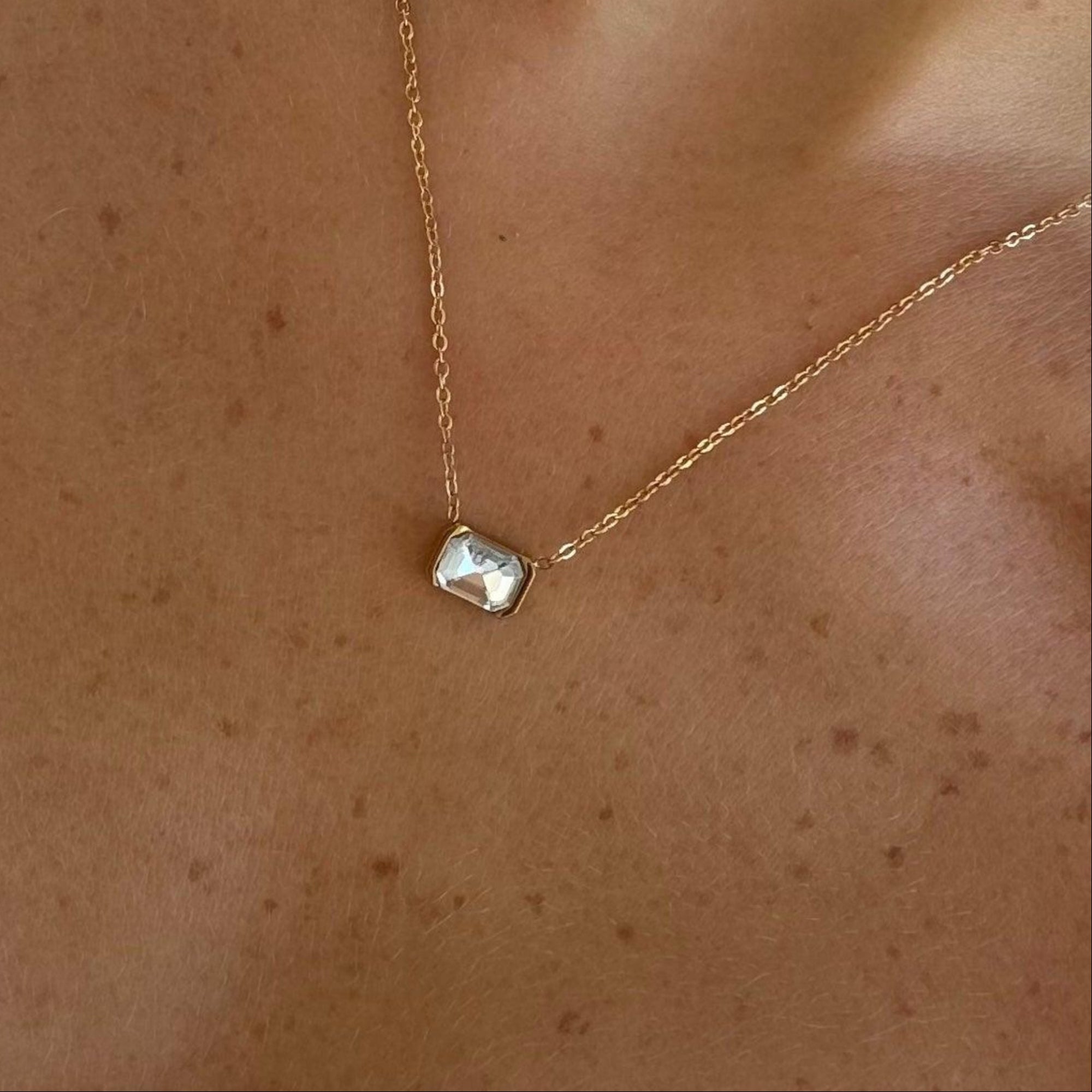 Dainty gold tone chain necklace with a clear rectangular bezel set stone worn at the collarbone