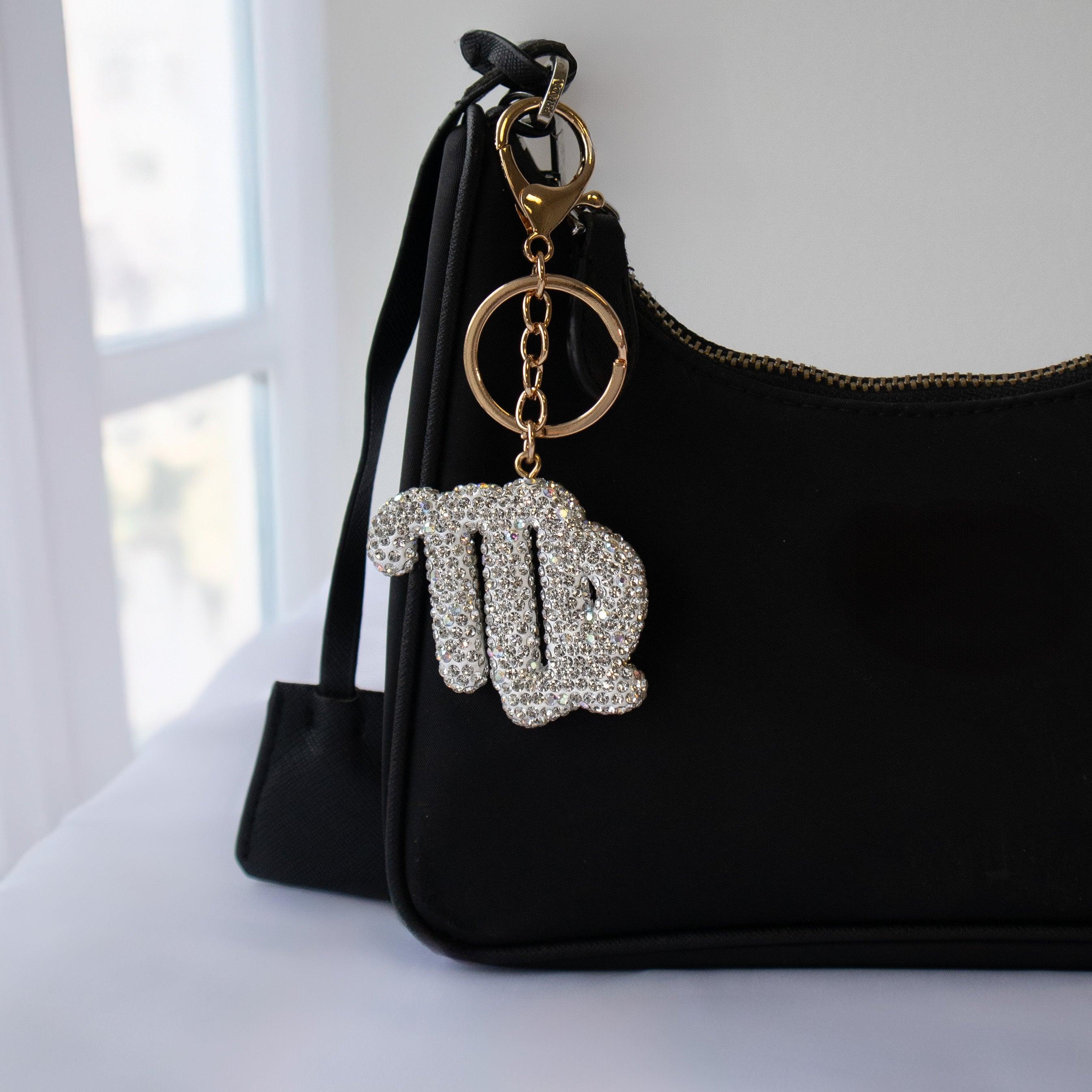 Sparkling Virgo zodiac symbol bag charm on a gold tone clasp hanging from a black shoulder bag